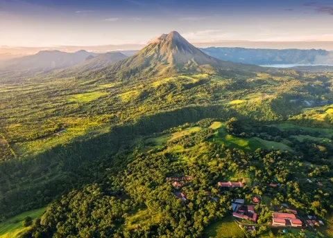 Aerial view of Arenal Volcano rising above lush green countryside with rolling hills, forests, and scattered buildings under soft morning light.