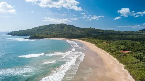 Aerial view of a sandy beach along Costa Rica’s Pacific coast, bordered by turquoise water and lush green hills.