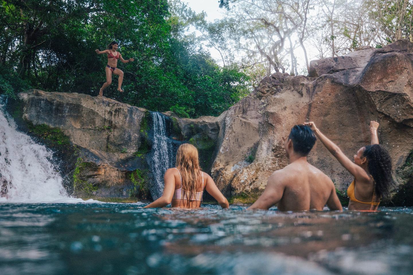Travelers swimming at the base of a rocky waterfall while one person jumps from a ledge into the natural pool in a forest setting.