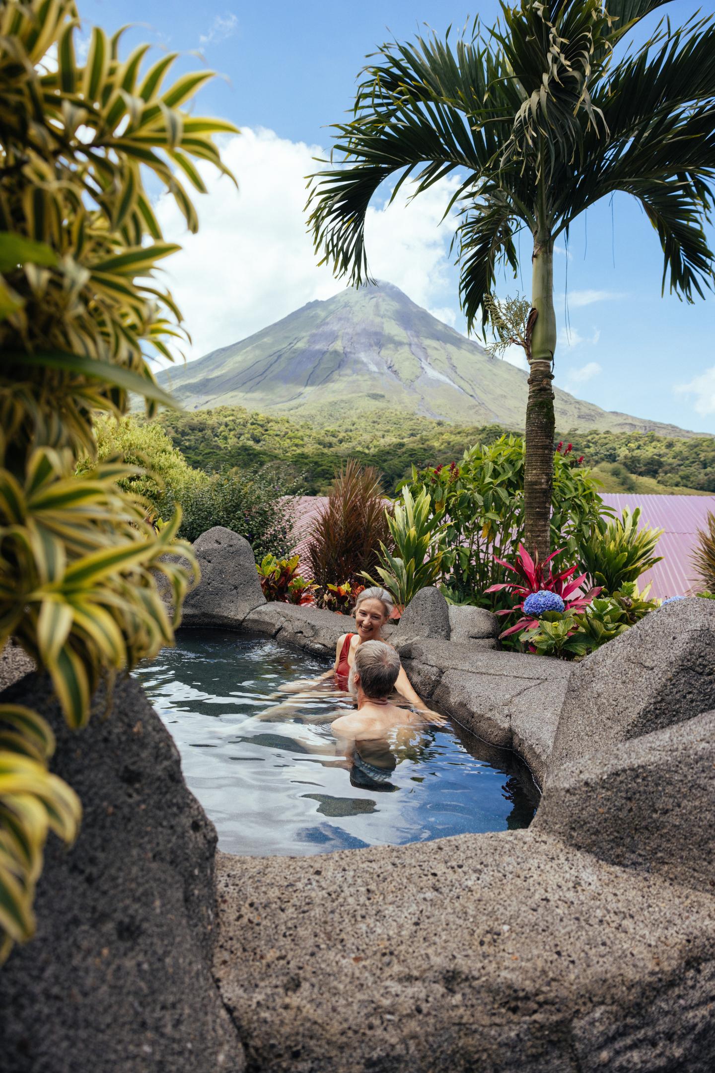 Two people relax in a stone hot spring pool surrounded by tropical plants, with Arenal Volcano in the background.