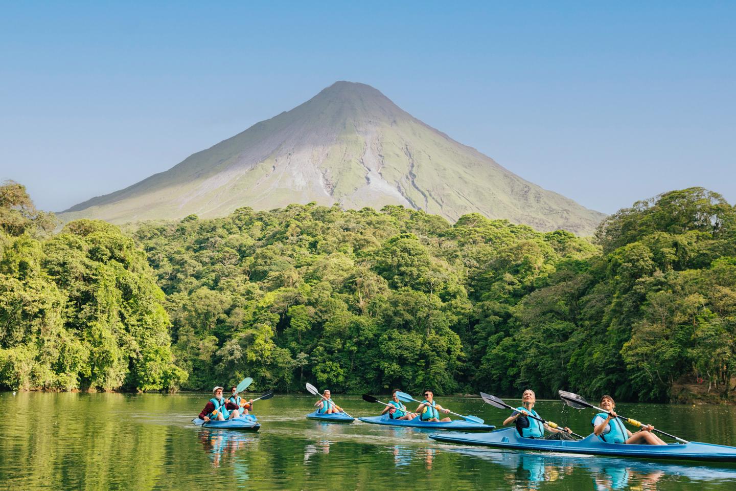 Group kayaking on a calm lake bordered by rainforest, with Arenal Volcano rising in the background.