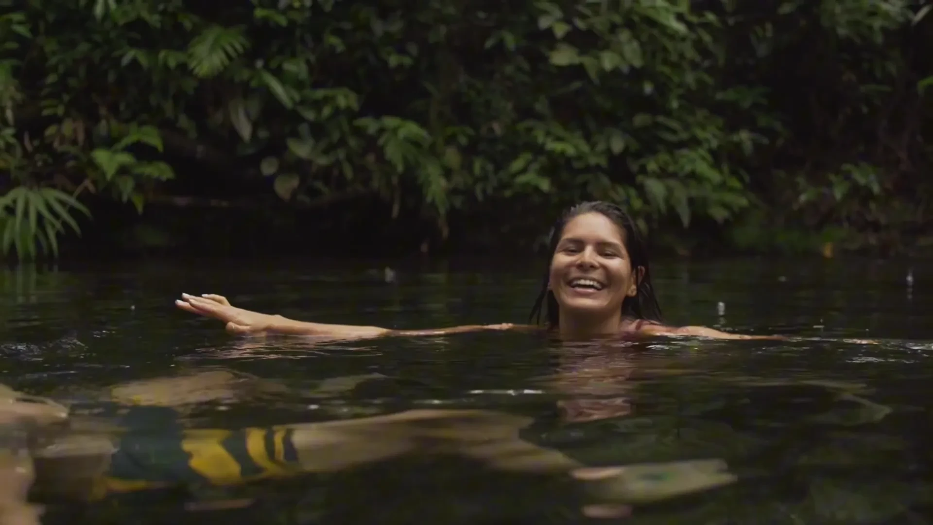 Woman laughing joyfully while swimming in a calm jungle river, surrounded by dense green foliage.